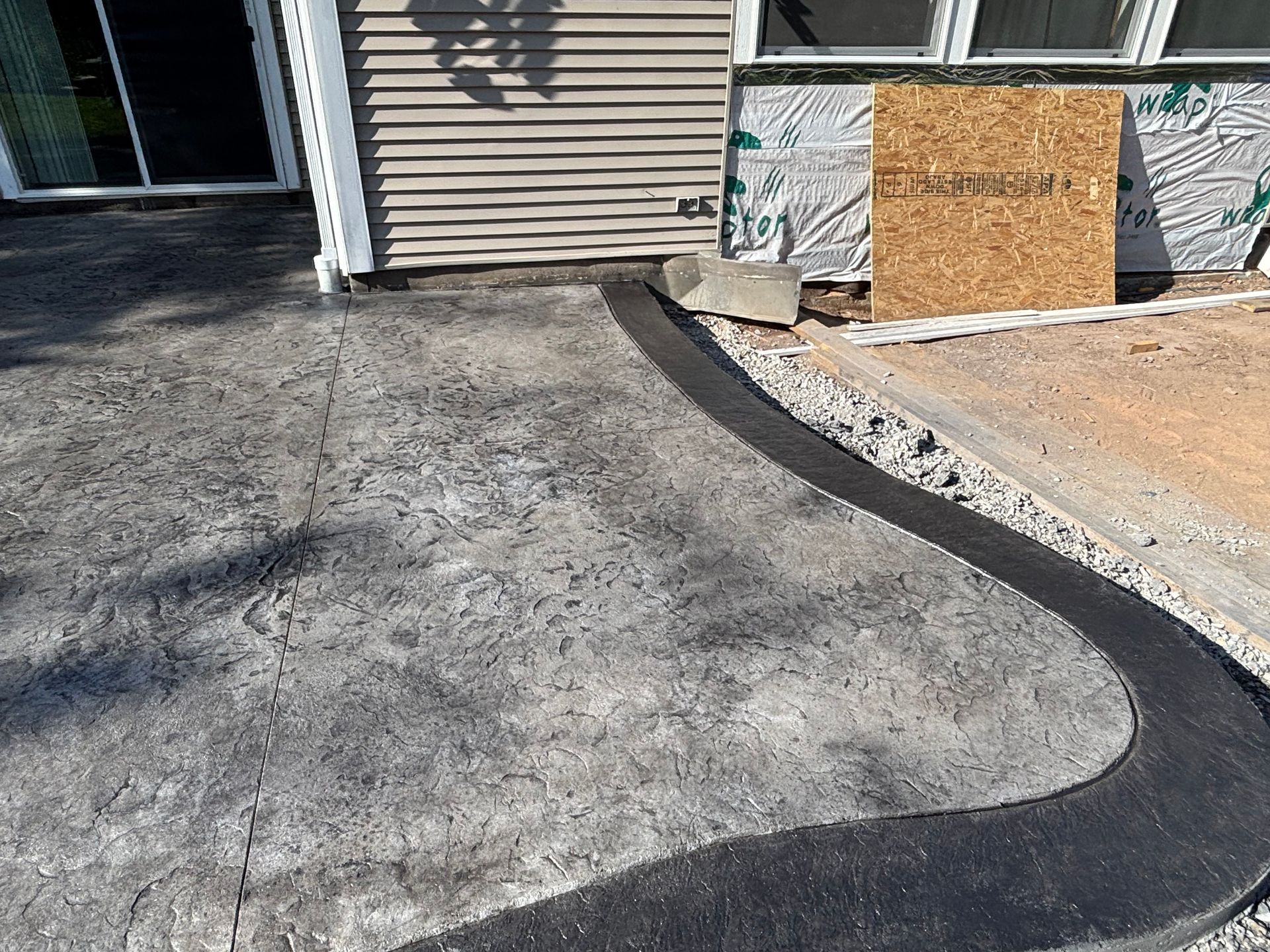 A textured grey concrete patio with a dark, smooth curved border next to a house under construction.