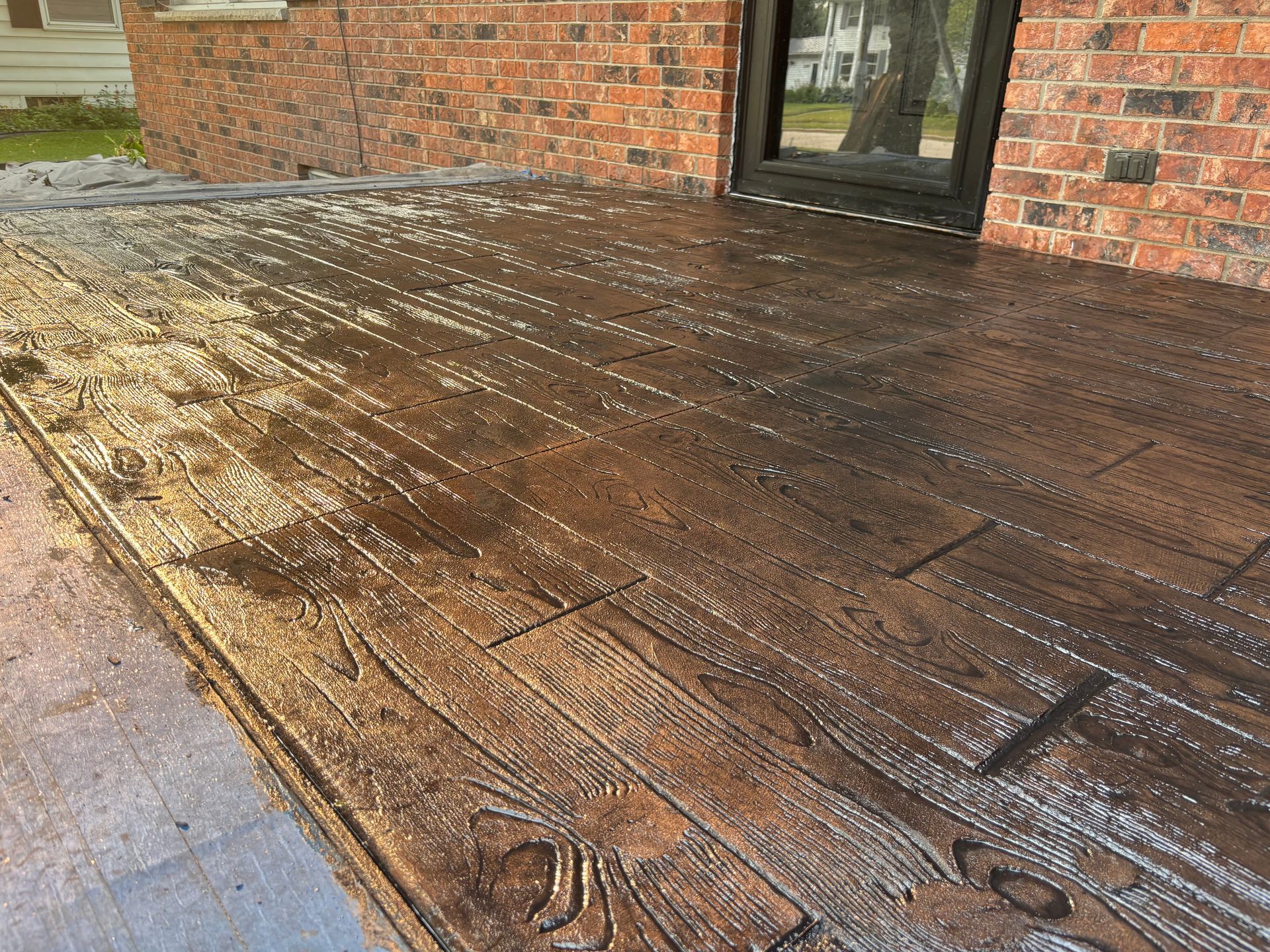 Stamped concrete patio textured to look like dark wood planks adjacent to a brick house wall.