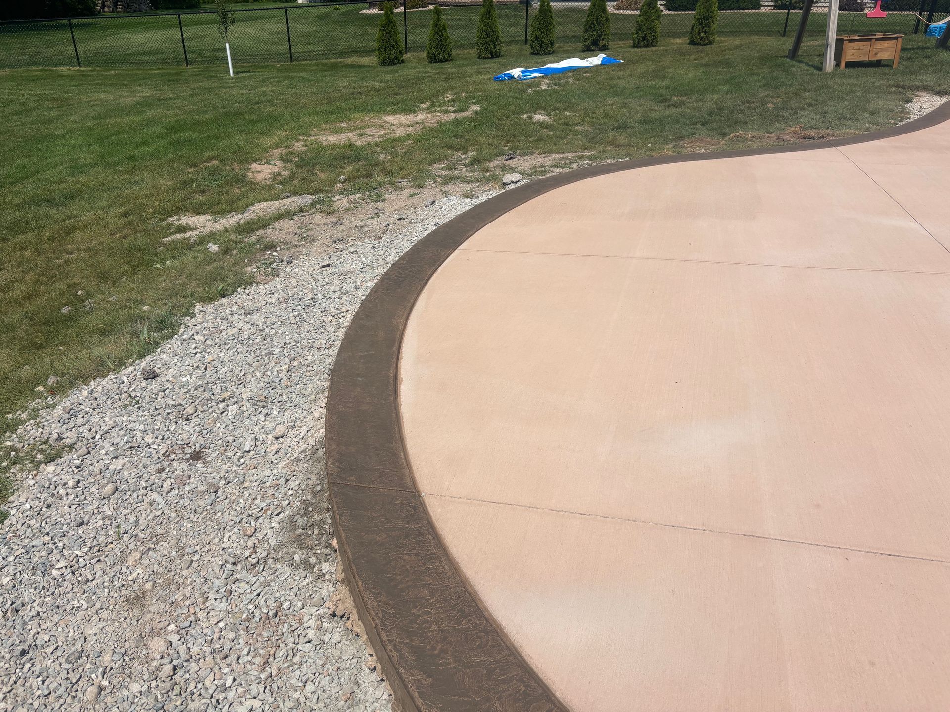 A curved concrete patio border with a dark textured edge sits adjacent to a gravel area and a grassy lawn.