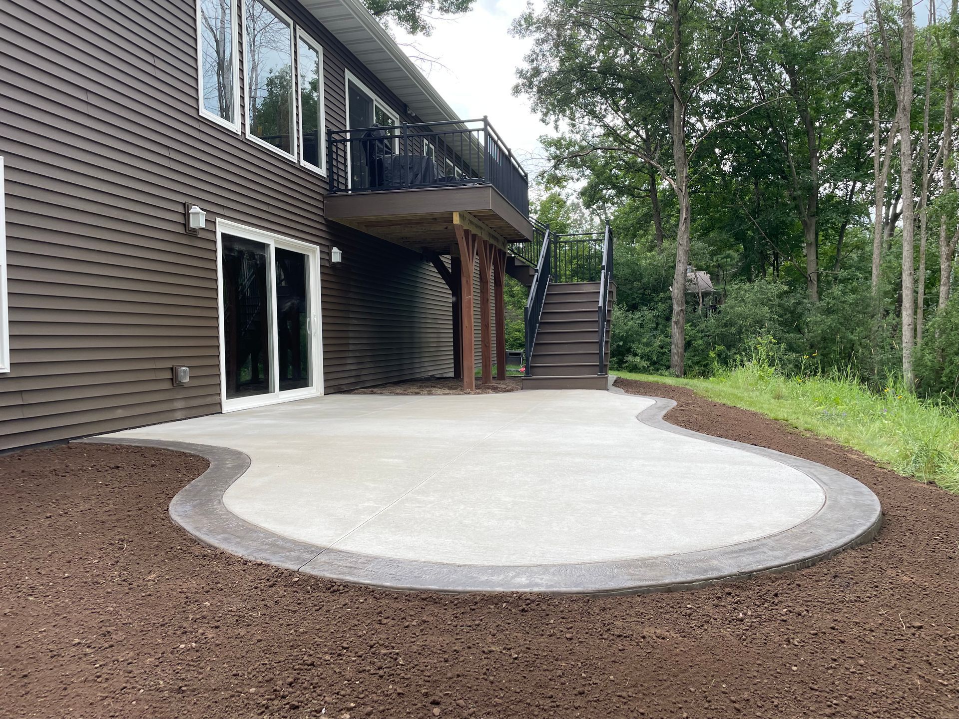 A modern brown house with a large concrete patio, a raised deck, and outdoor stairs, surrounded by fresh landscaping mulch.