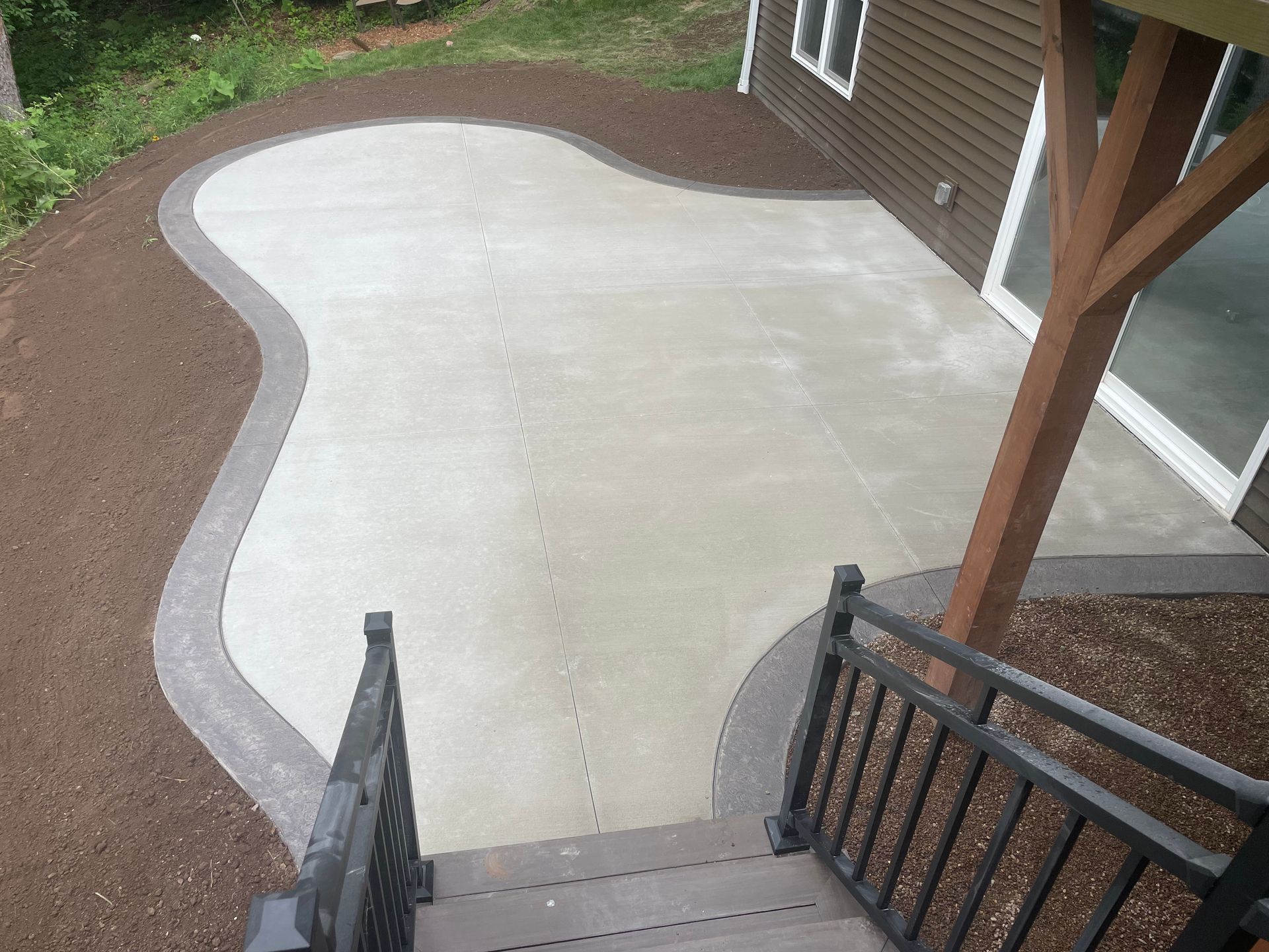 A curved light gray concrete patio with a dark border, bordered by brown mulch, viewed from a wooden deck staircase.