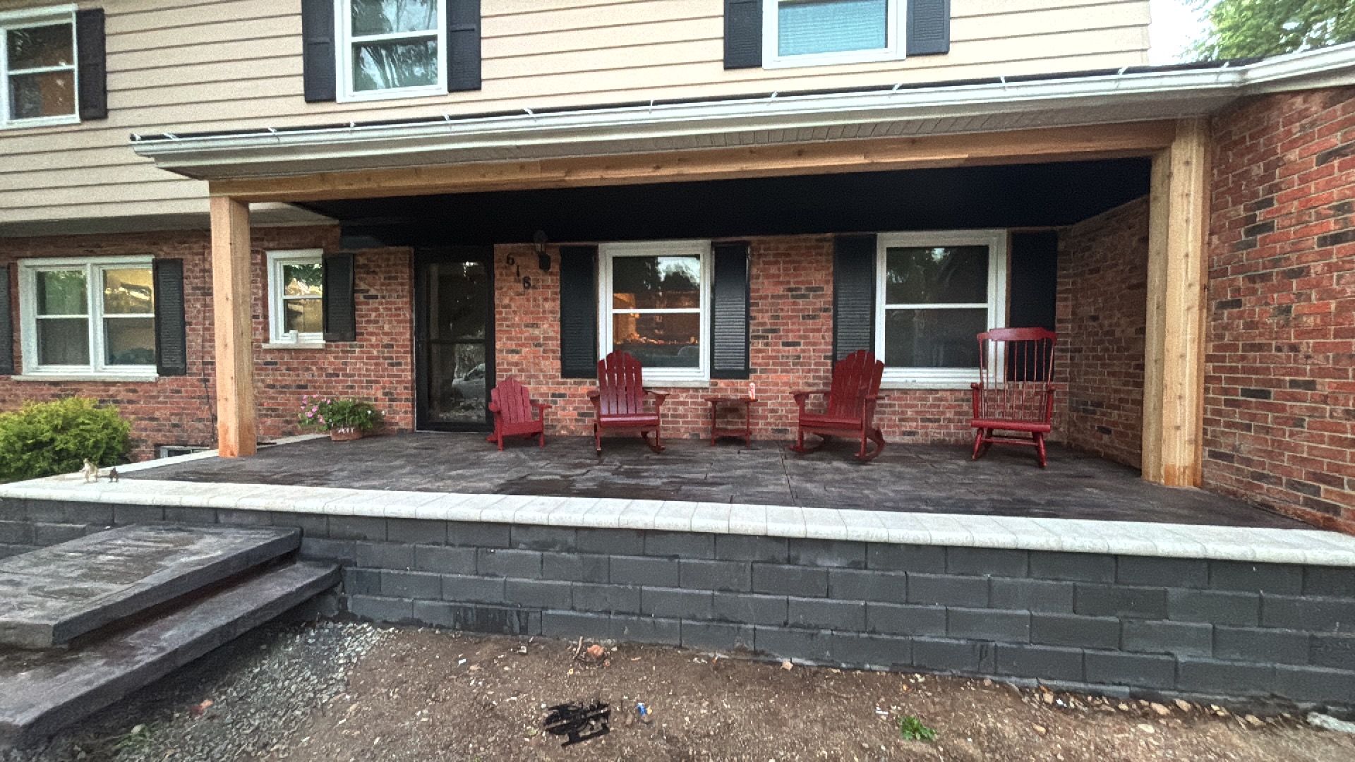 A covered brick porch with a grey stone base, featuring four red chairs, black shutters, and light siding above.