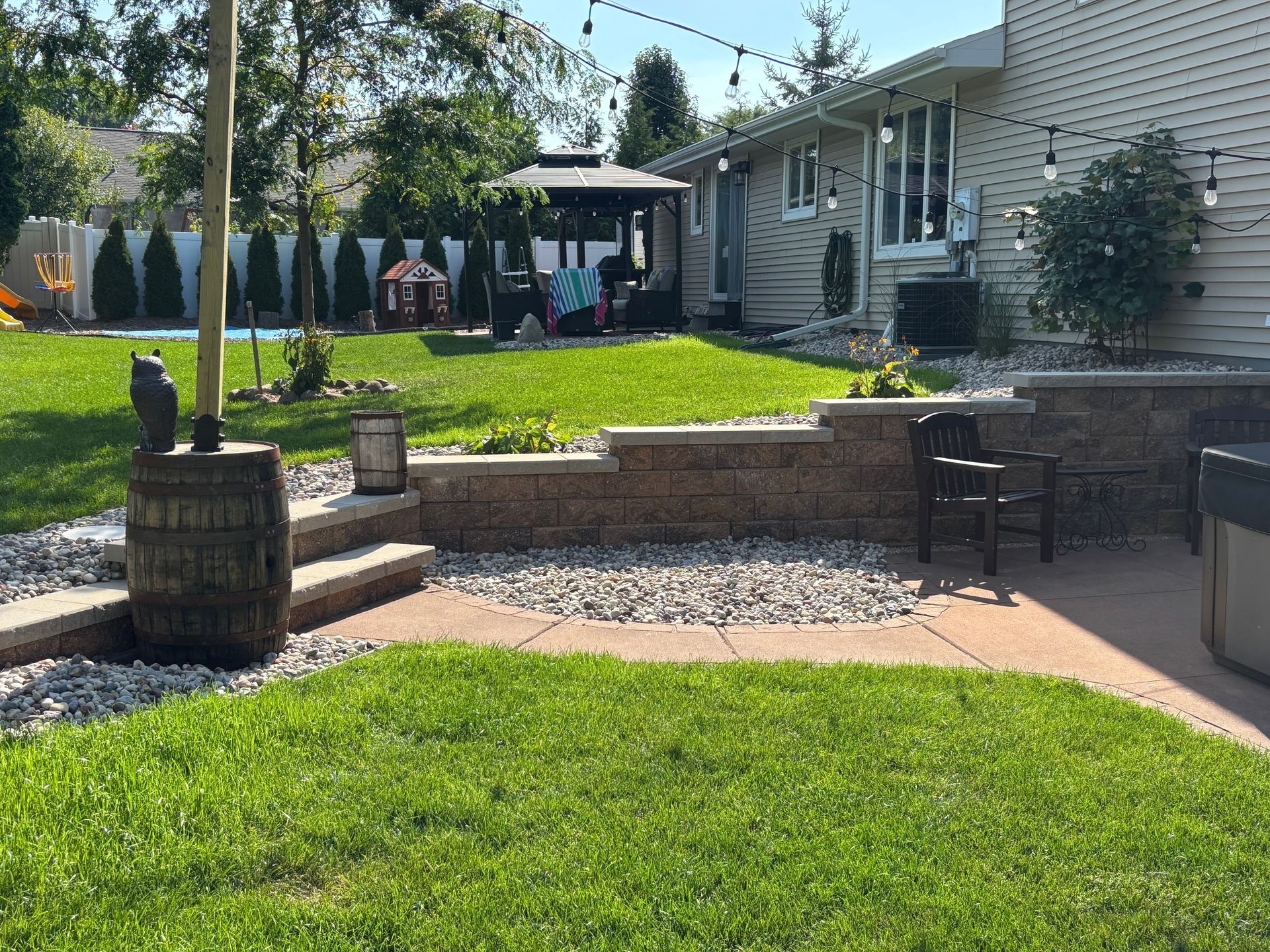 A backyard featuring a stone retaining wall, a paved patio, a wooden barrel, string lights, and a gazebo on the lawn.