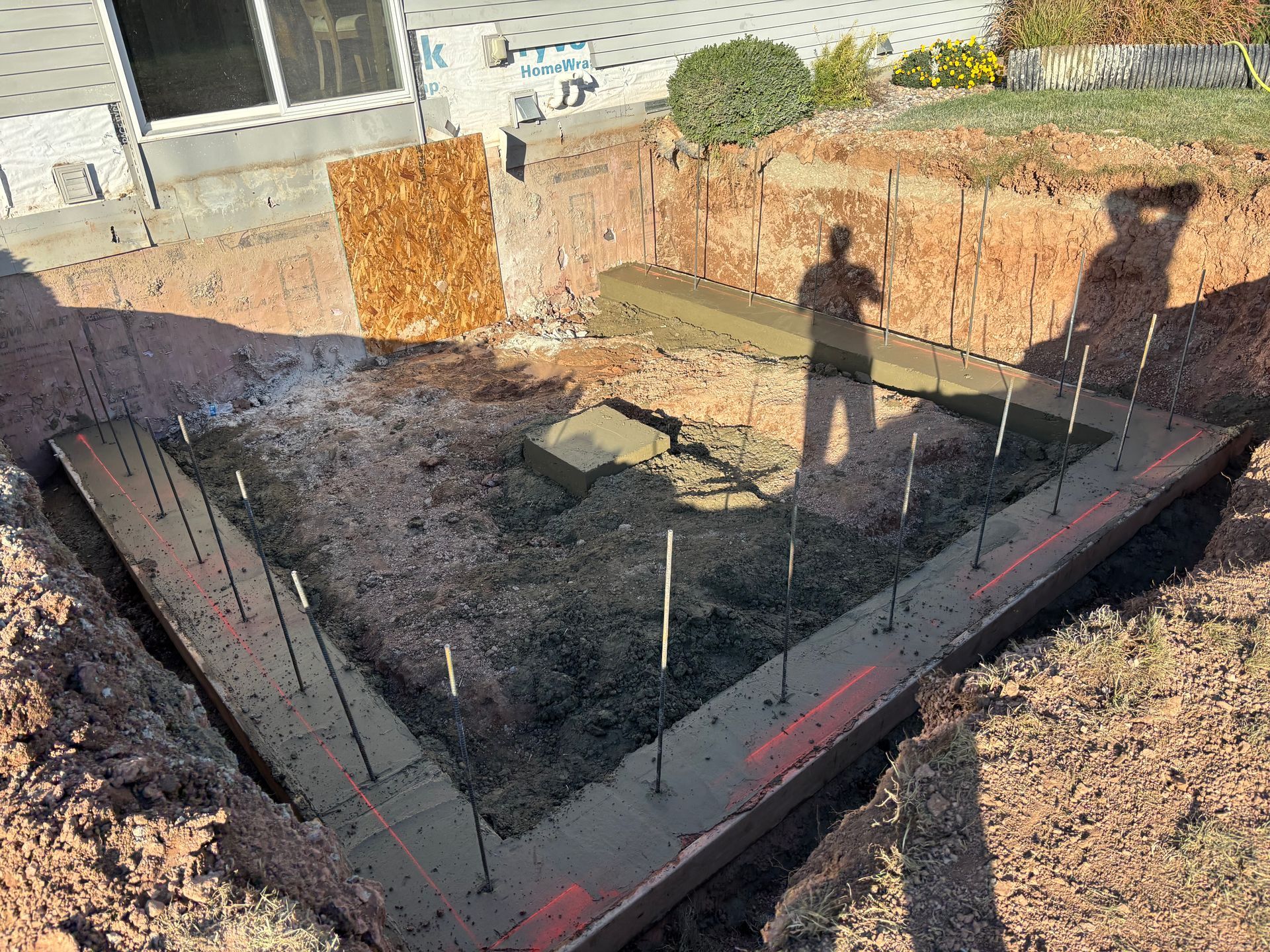 A freshly poured concrete foundation for a building addition sits in an excavated dirt pit next to a house exterior.