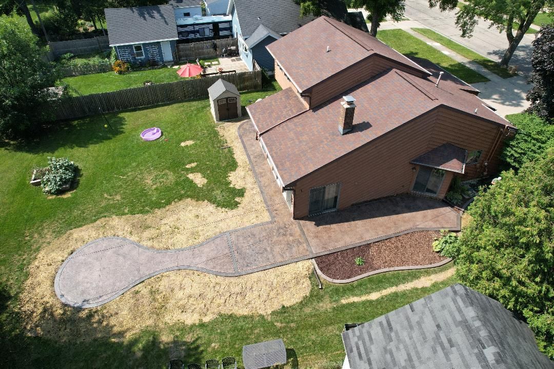 Aerial view of a brown house with a stamped concrete patio and walkway extending into a backyard with patches of dirt.