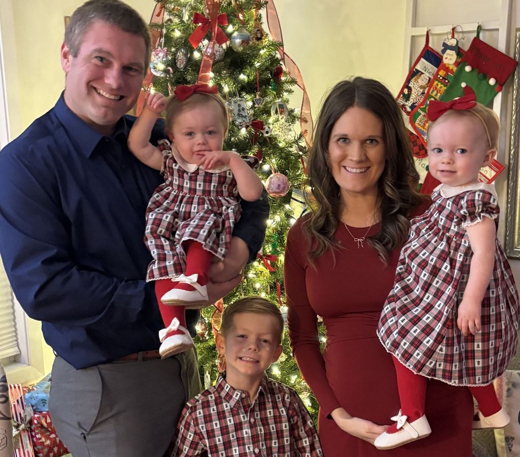 A family of five posing in front of a decorated Christmas tree, all wearing coordinating red and plaid holiday outfits.