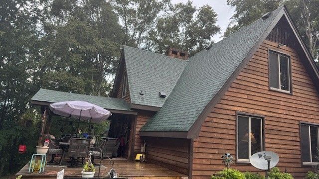Two roofers in orange and yellow hardhats installing asphalt shingles on a rooftop. One is hammering while the other kneels.