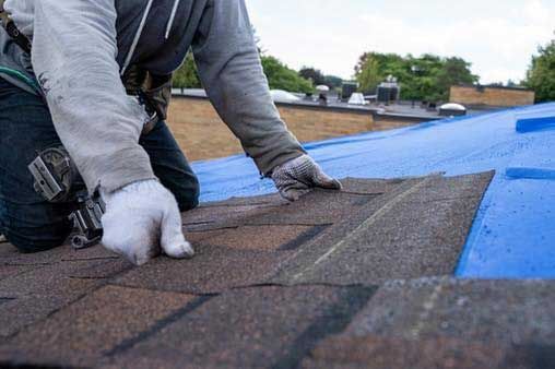 A roofer kneels on a blue tarp-covered roof, installing brown asphalt shingles. They wear work gloves and a safety harness.