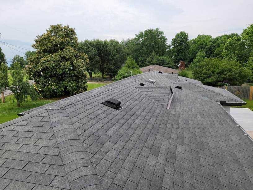 Gray asphalt shingle roof of a house, viewed from above, with vents and a green lawn and trees in the background.