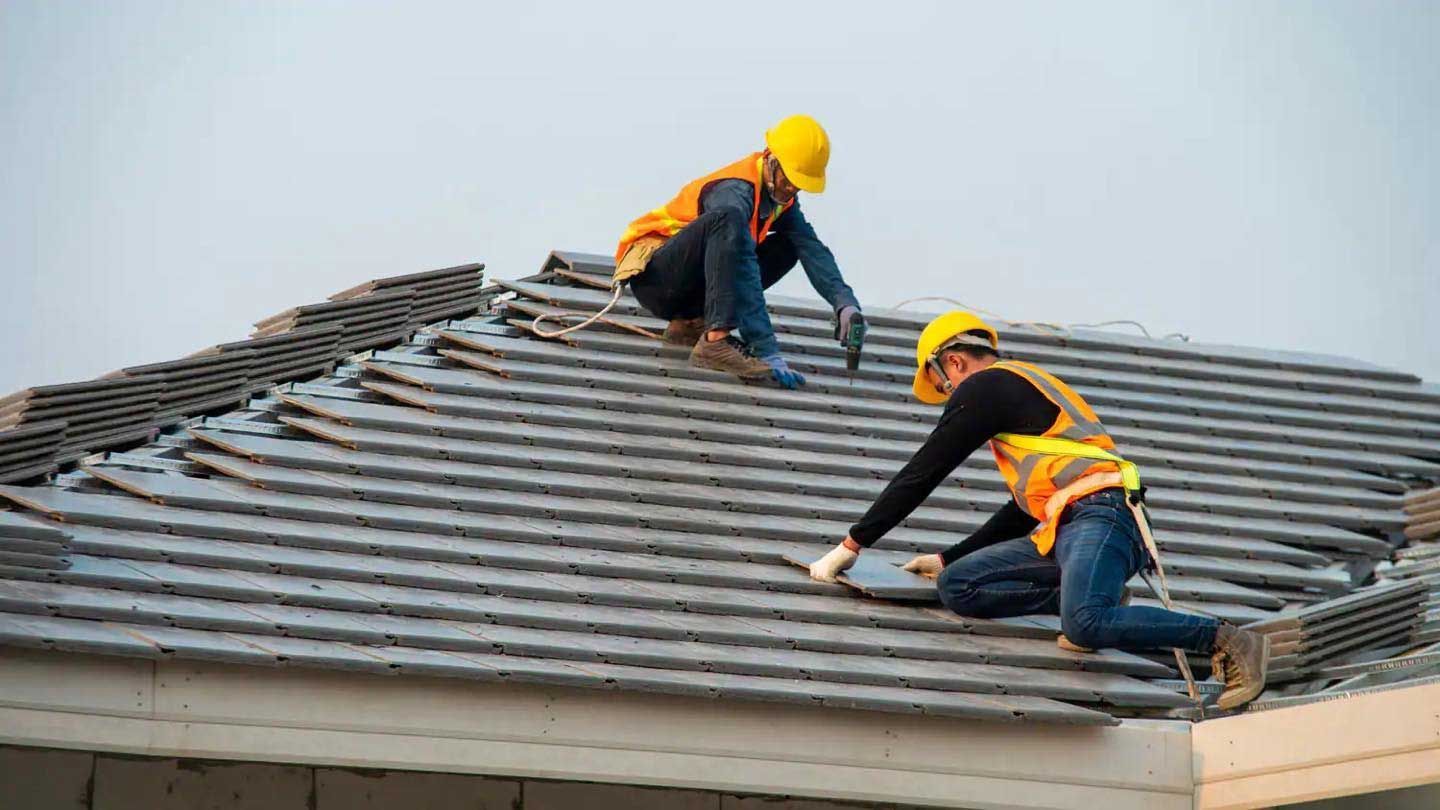 Two roofers in safety gear installing tiles on a roof. One is kneeling and placing a tile, the other is using a power drill.