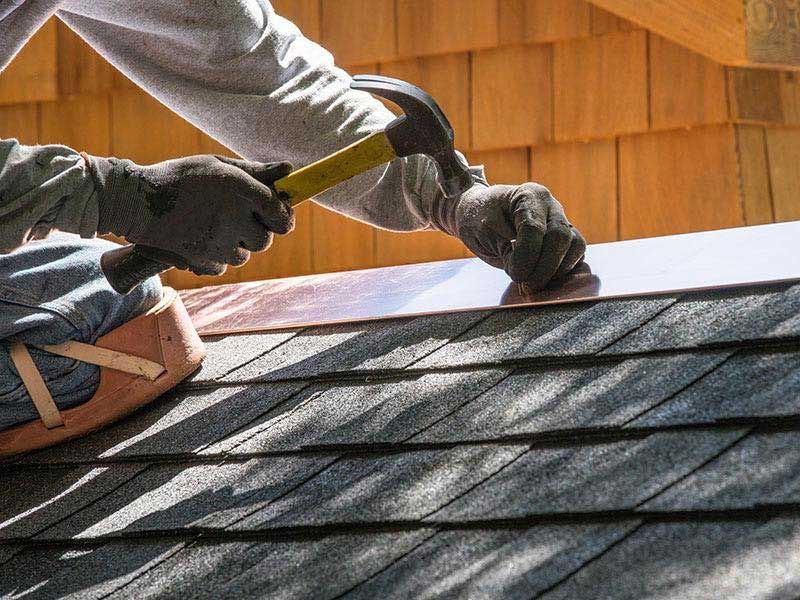 Person hammering copper flashing onto a roof, wearing gloves, with a wood shingle background.