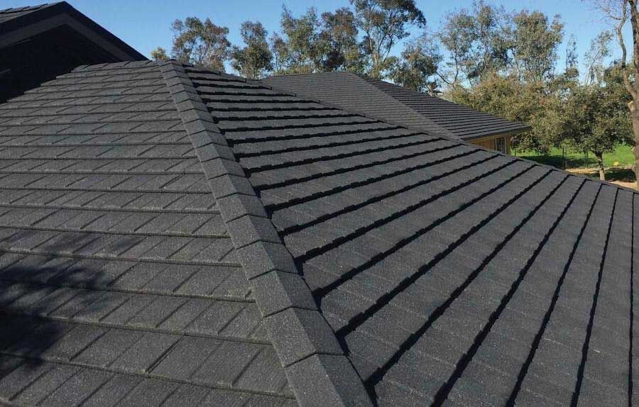 Close-up of a black asphalt shingle roof on a house, with blue sky and trees in the background.