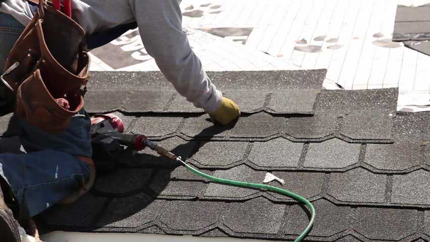 A roofer installs shingles on a roof, using a nail gun. The work is outdoors in daylight.