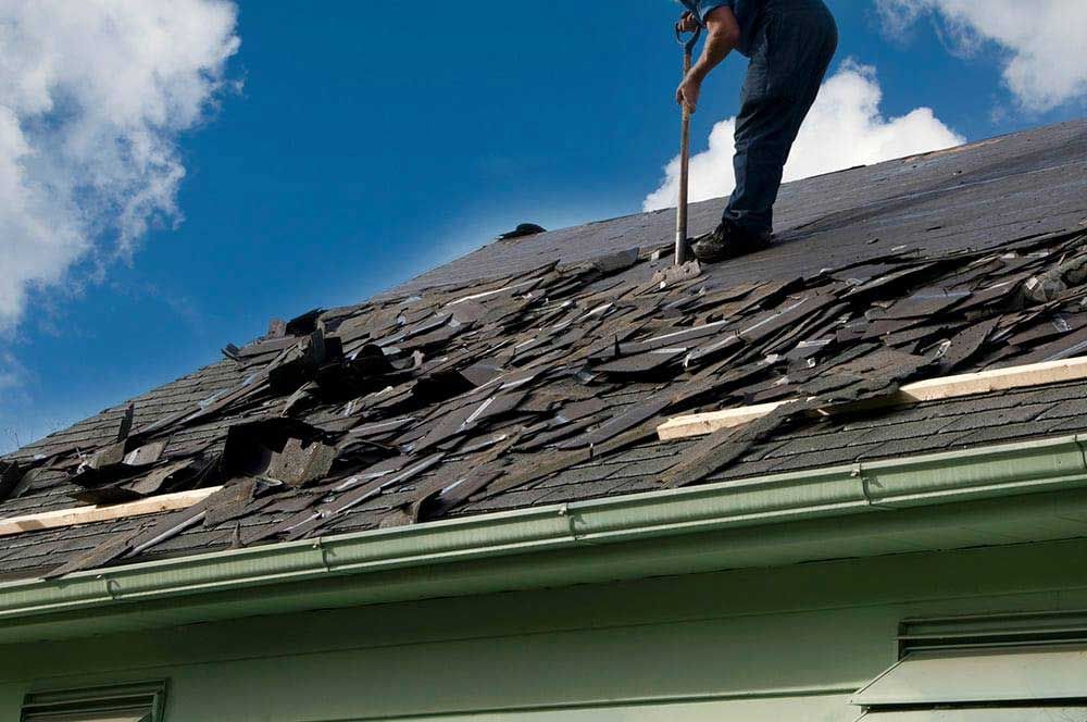 Person removing damaged asphalt shingles from a house roof under a blue sky with clouds.