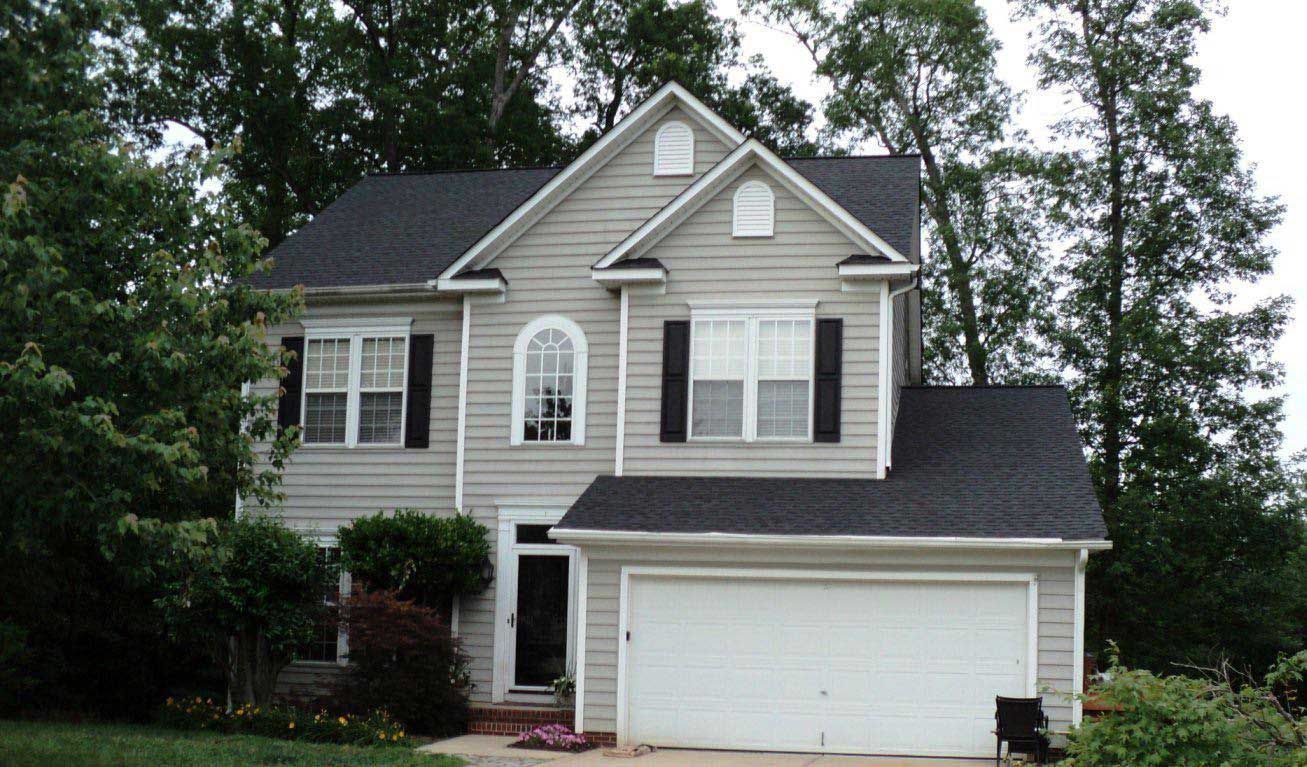 Two-story house with light gray siding, a dark roof, and a two-car garage; surrounded by trees and greenery.