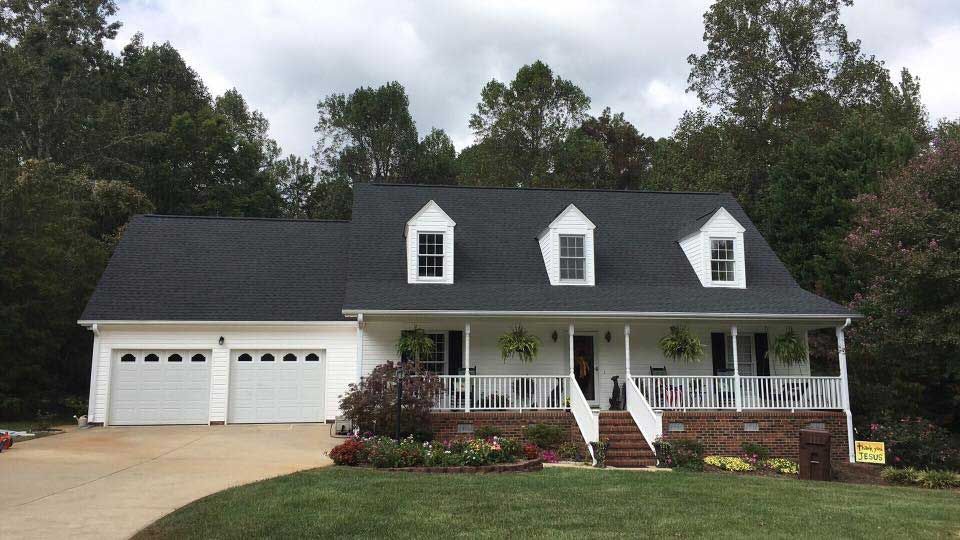 White house with black roof, two-car garage, porch, and dormers. Landscaping and trees surround the home.