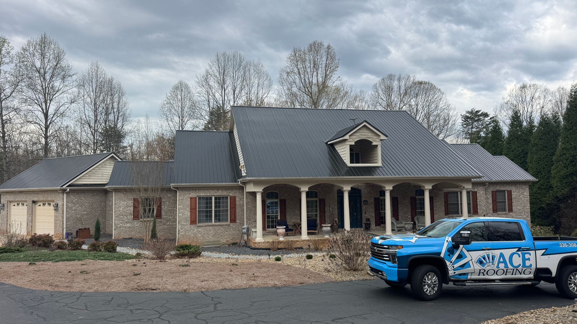 A single-story brick house with a dark metal roof and front porch, with a blue company truck parked in the driveway.