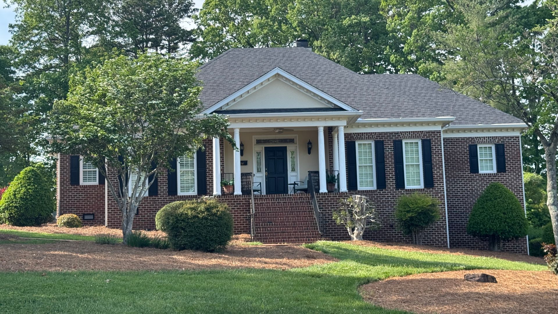 Brick ranch house with white columns, black shutters, and a front porch surrounded by trees and lawn