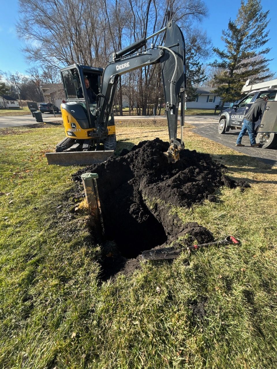Excavator digging a large hole in a grassy yard with dirt piled beside it, under a blue sky.
