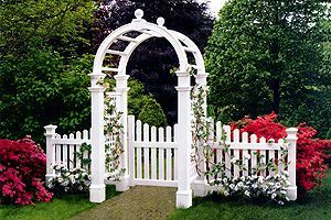 A white archway with a gate in the middle of a garden.