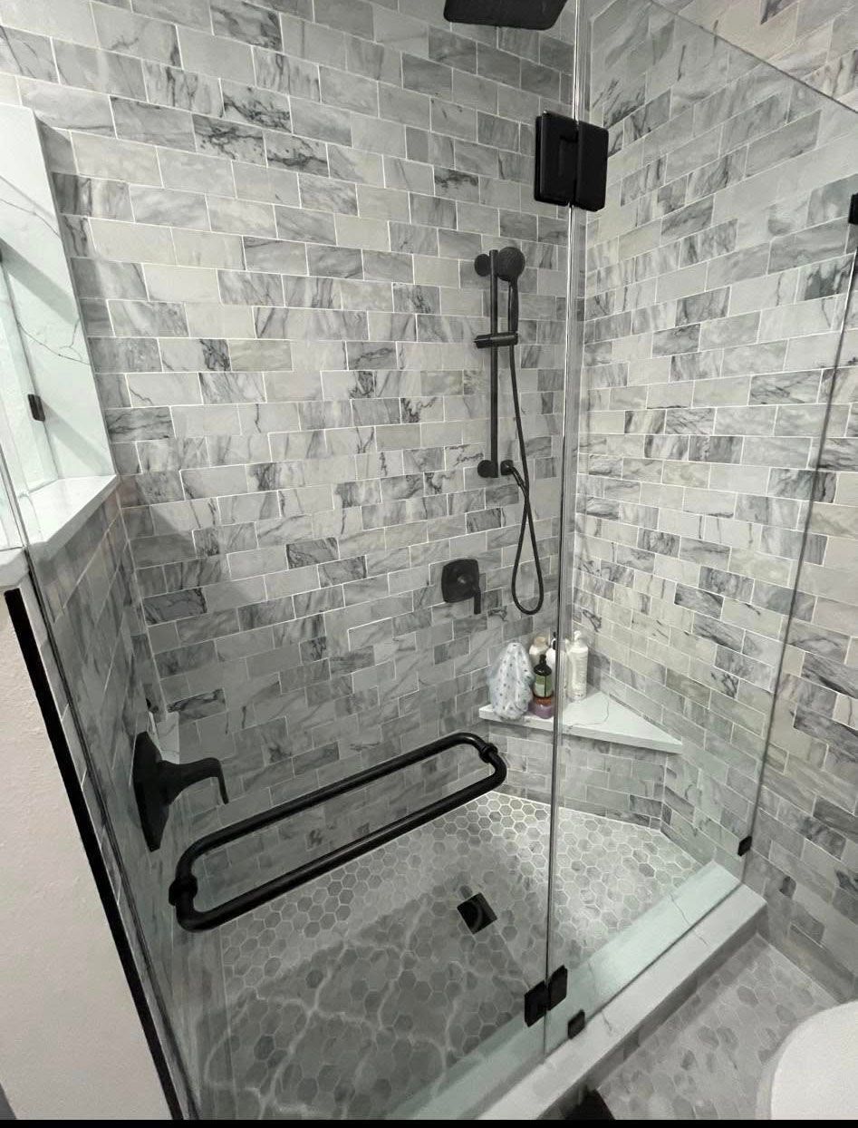 Gray tiled shower with glass door, black fixtures, and built-in corner seat.