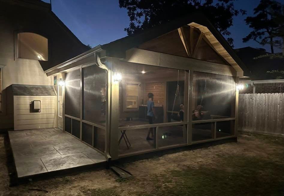 Screened-in porch lit at night; attached to a house. A person stands inside; backyard with fence.