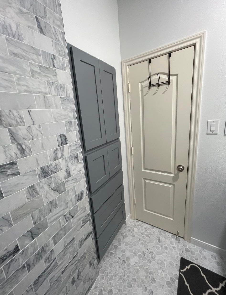 Gray cabinet and white door in a bathroom with patterned floor and stone-look wall.