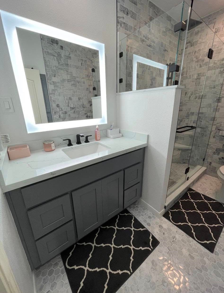 Modern bathroom with grey vanity, lighted mirror, and patterned tile floor. Glass shower.
