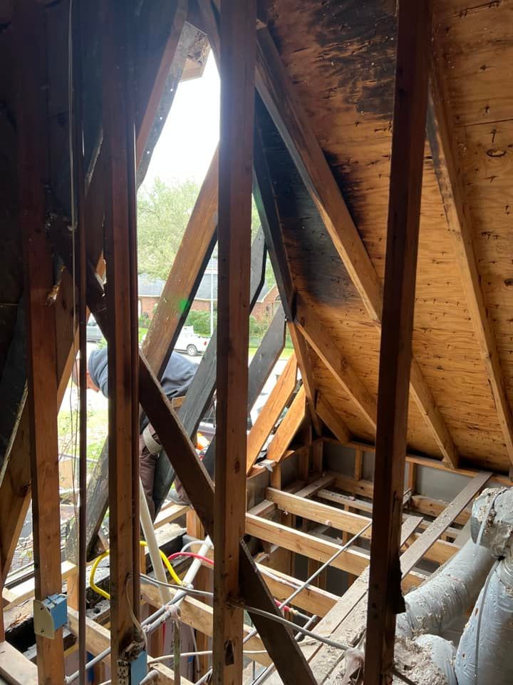Interior view of an attic with exposed wooden rafters. A person works amid exposed framing, and daylight streams in.