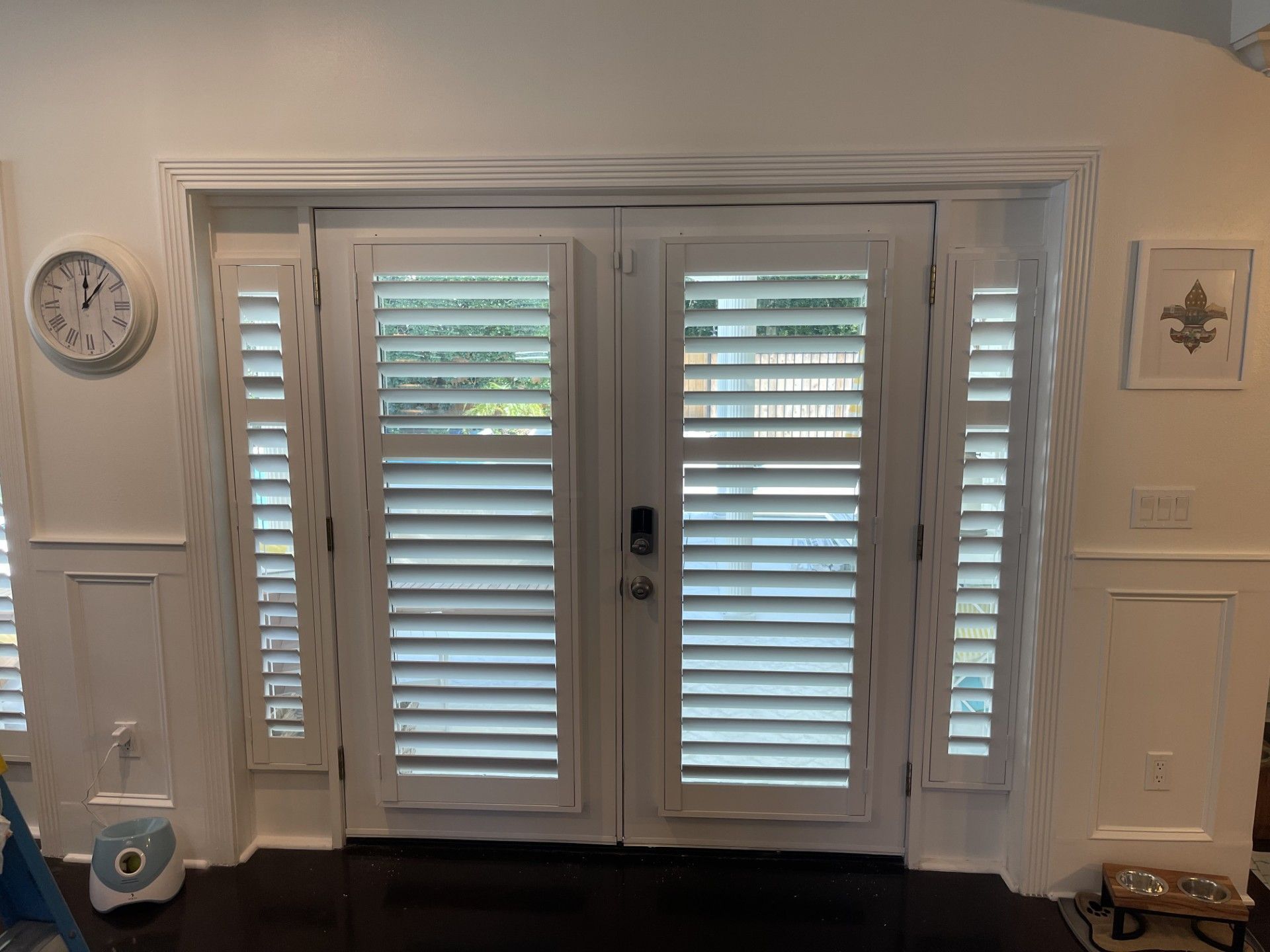 White shuttered double doors flanked by sidelights, with a clock and wall decor nearby.