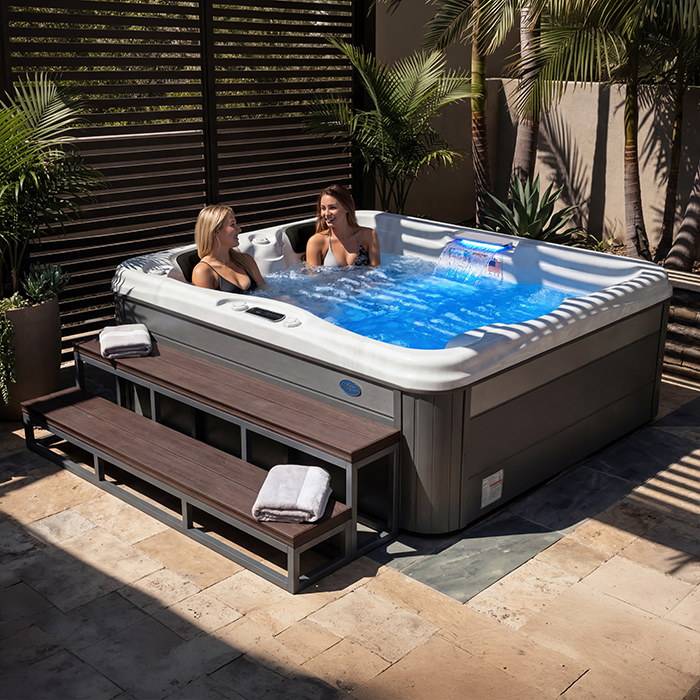 Two people relaxing in a blue hot tub on a sunlit wooden patio with plants and slatted privacy screens