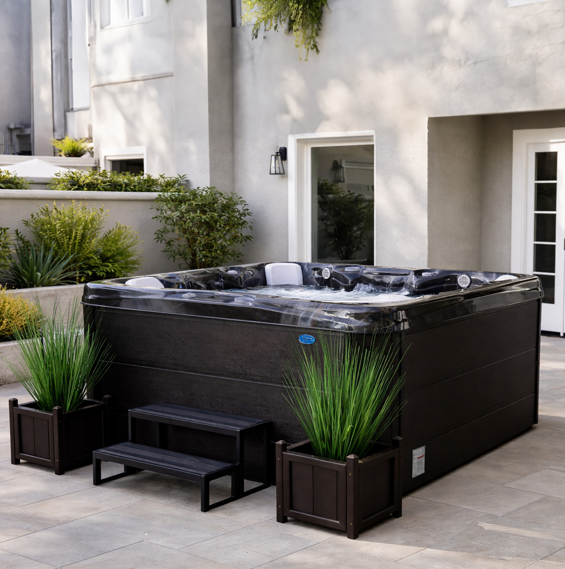 Black outdoor hot tub on a patio beside a white house, with small planters and a bench nearby
