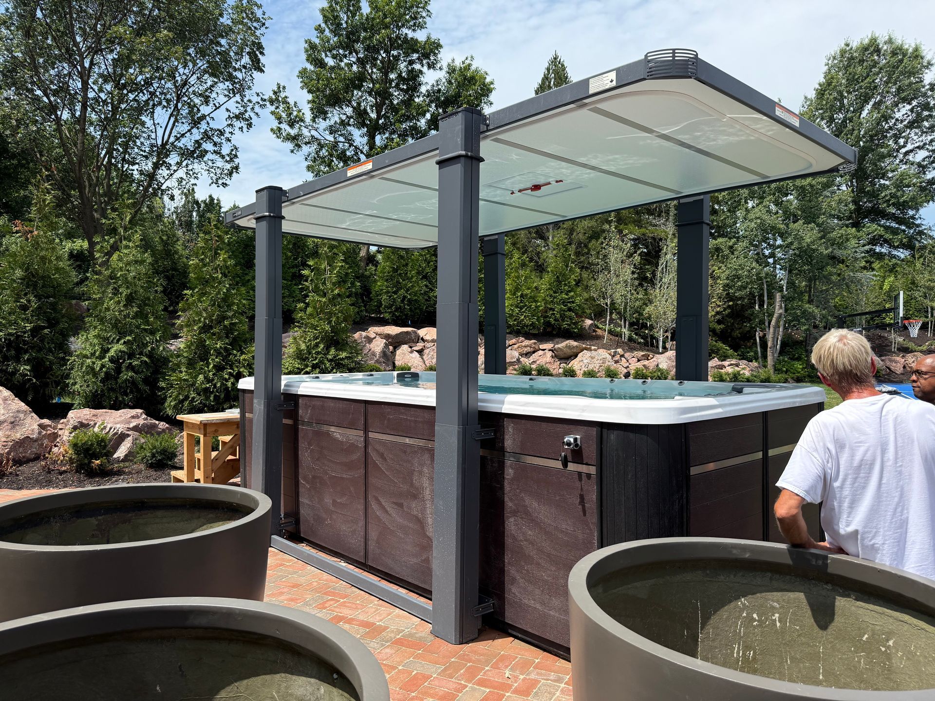 Hot tub with a canopy in a backyard setting. Person is by the tub, large planters in the foreground.
