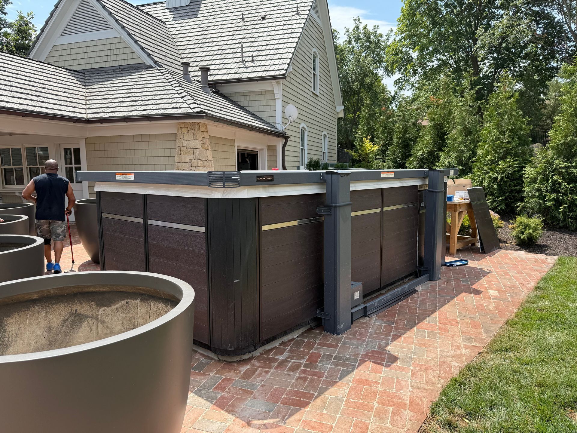 A hot tub with a cover and a person working on it, set on a brick patio next to a house.