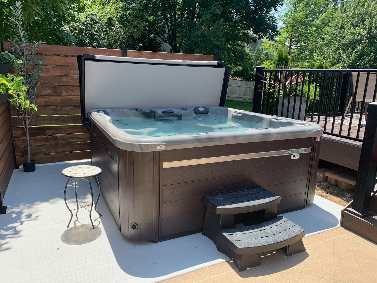 Hot tub with partially open cover on a concrete patio, next to a small stool and steps. Wooden fence and deck in the background.