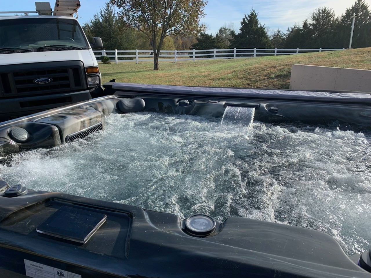 Hot tub with bubbling water, a small waterfall, and a white van in the background, set outdoors on a sunny day.