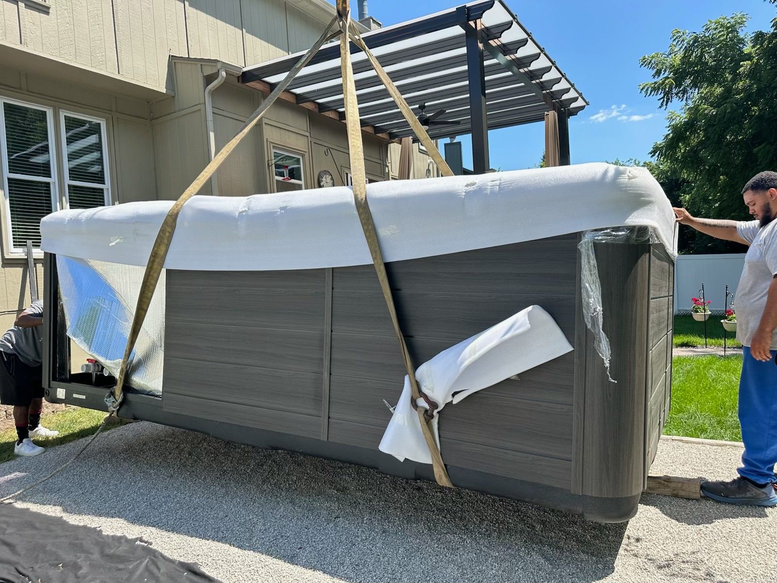 Two men lifting a hot tub with straps; one on ground, the other guiding. Outdoors, near a house with a pergola.