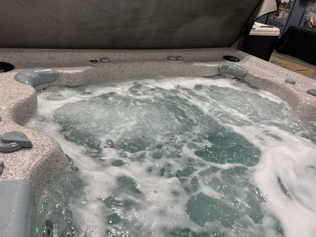 Bubbling hot tub with jets creating foam. Beige and gray tub edge, partially open cover.