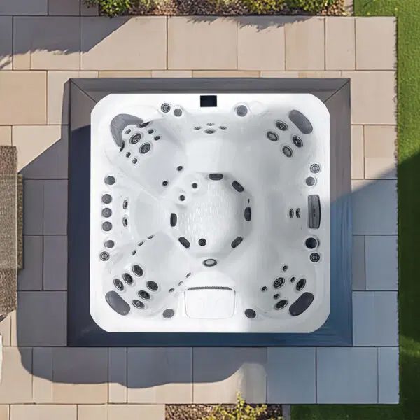 An overhead view of a square, white hot tub with gray headrests and jets, installed on a light-colored stone patio.