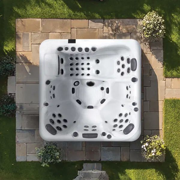 A top-down view of a white, square hot tub centered on a stone patio surrounded by grass.
