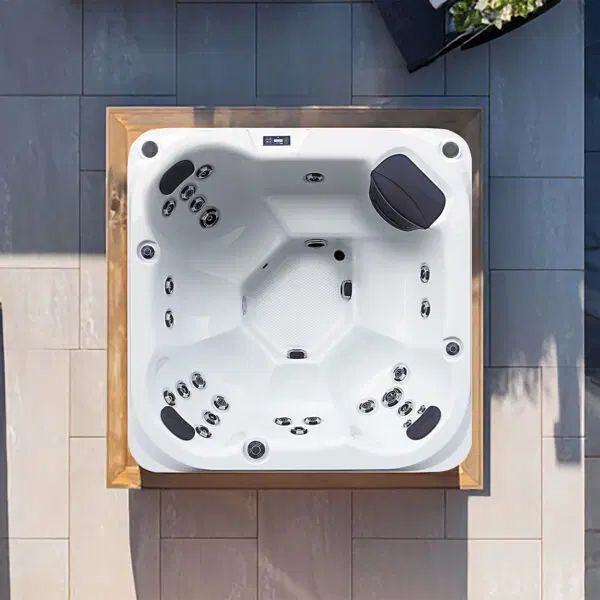 Overhead view of a square, white hot tub with dark grey headrests, set in a light wooden frame on a stone patio.