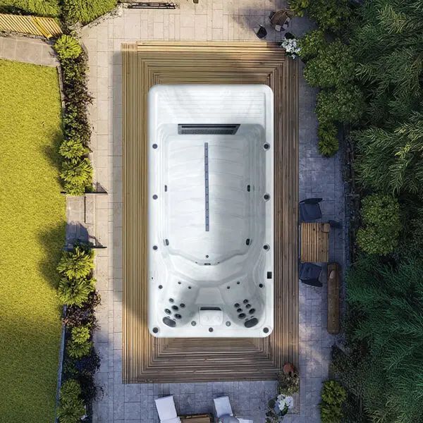 A top-down view of a rectangular, white swim spa set into a wooden deck in a lush, green outdoor garden setting.