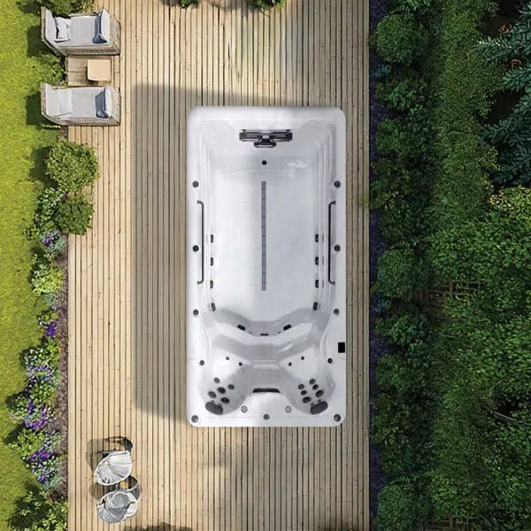 A white rectangular swim spa sits on a light wooden deck next to a lush garden, viewed from directly above.