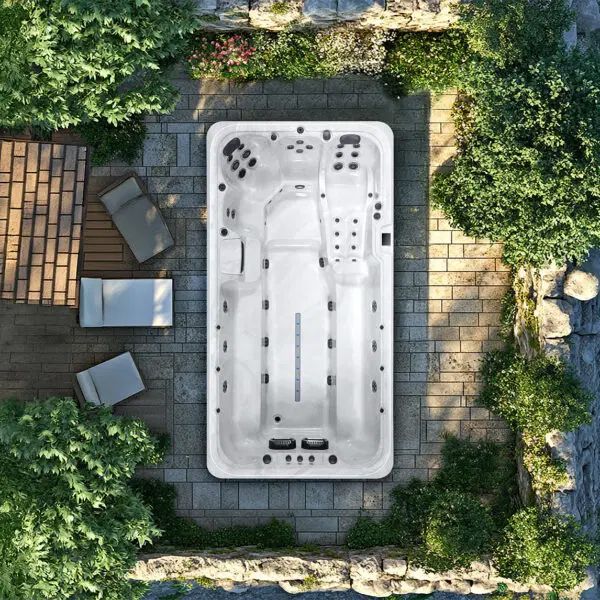 An overhead view of a white rectangular swim spa centered on a stone patio, surrounded by lush green foliage and furniture.