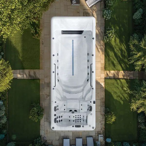 An overhead view of a white, rectangular swim spa set in a stone patio surrounded by green lawn and trees.