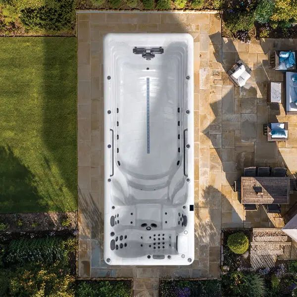 An aerial view of a rectangular, white swim spa set into a stone patio, flanked by a lawn and outdoor lounge furniture.