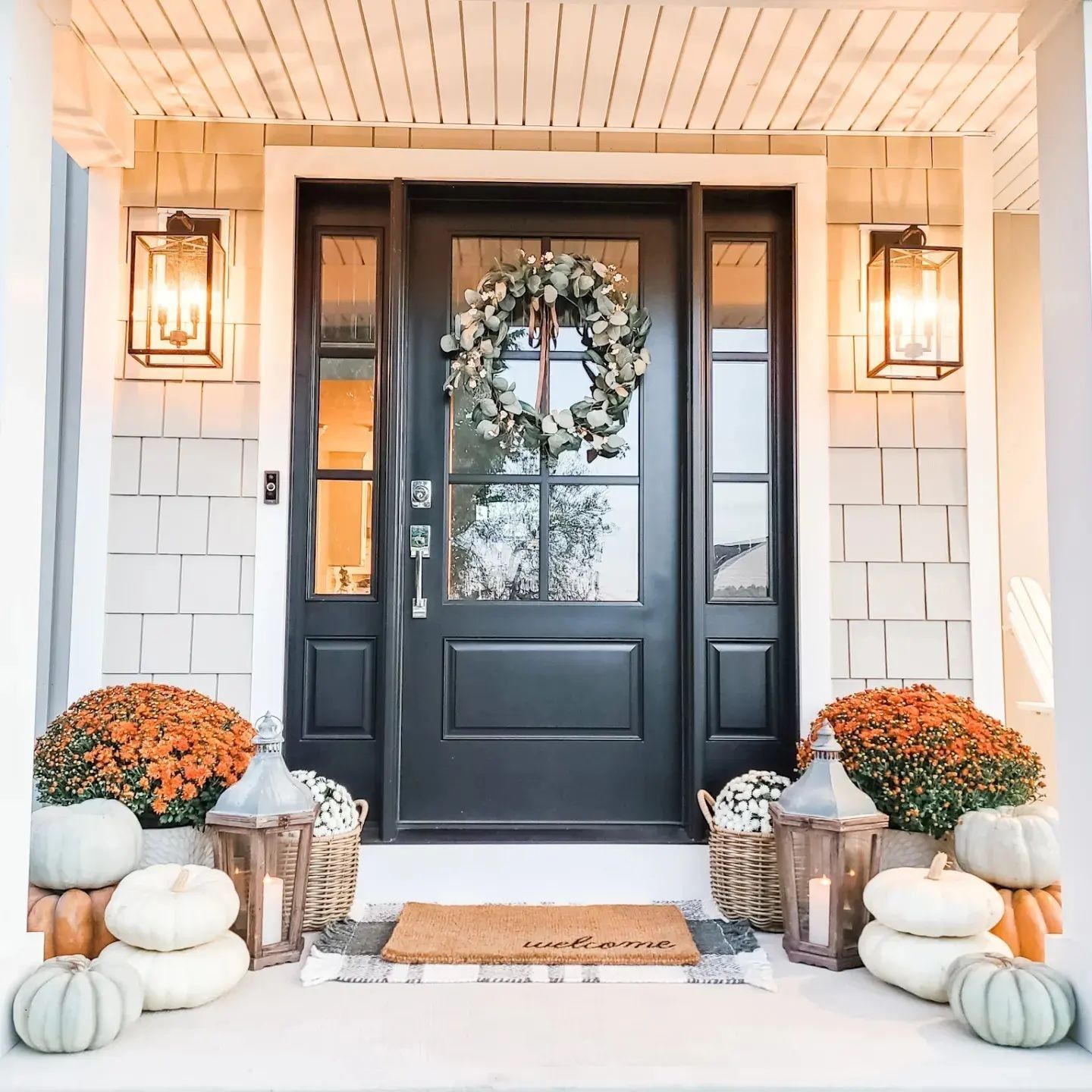 A black door with a wreath on it is surrounded by pumpkins and flowers