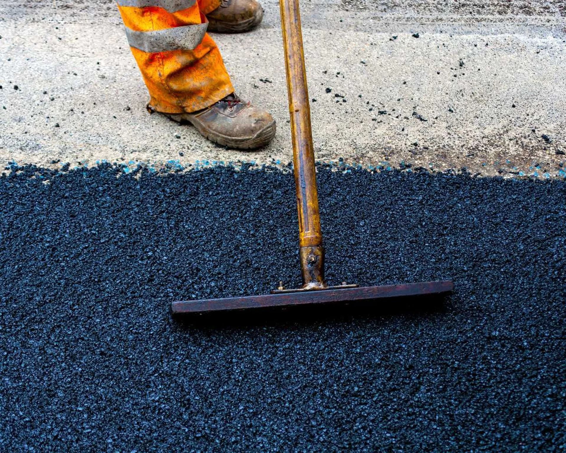 A worker levels fresh black asphalt with a rake beside orange safety pants and boots.