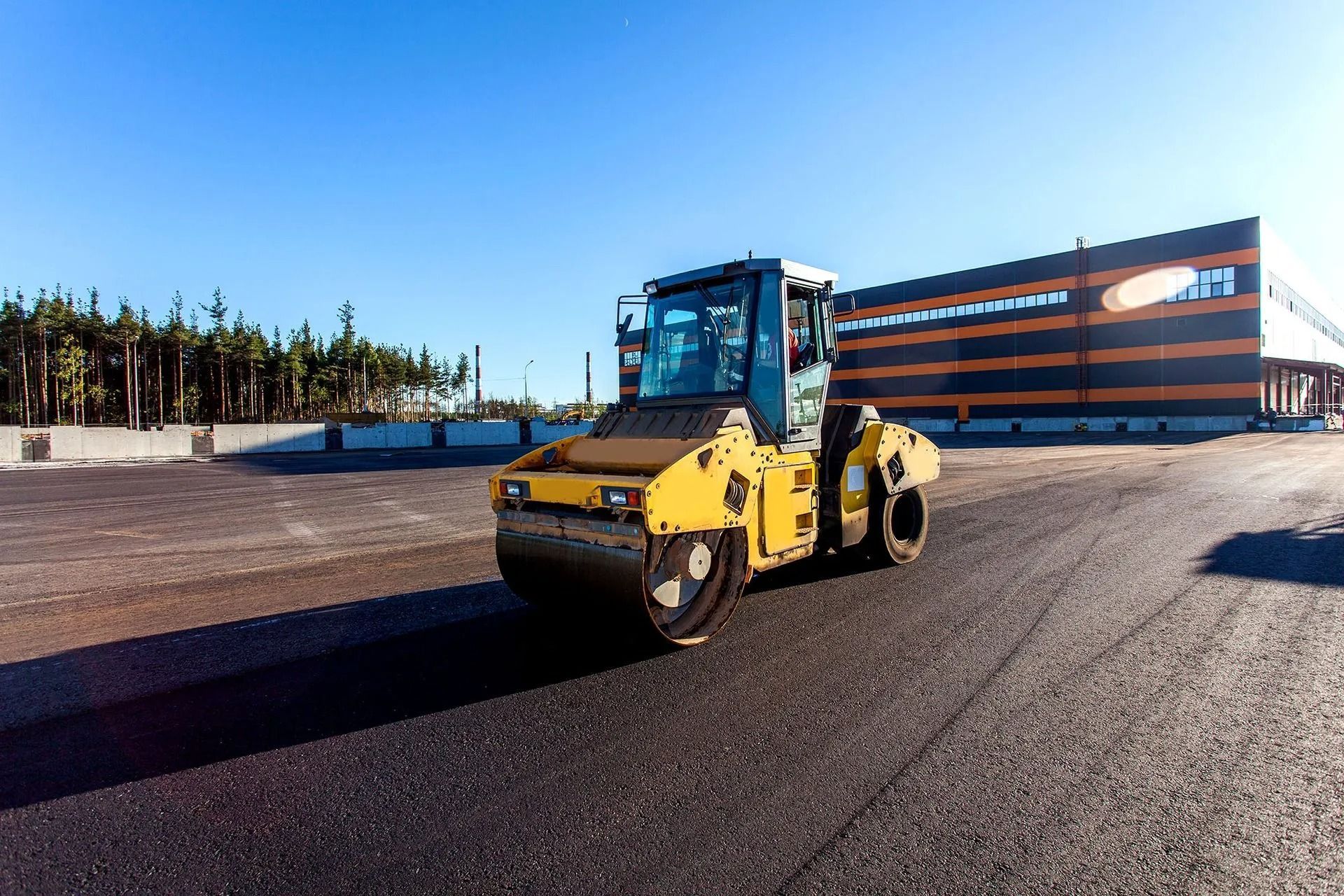 Yellow road roller on a paved surface near a modern building under a clear sky
