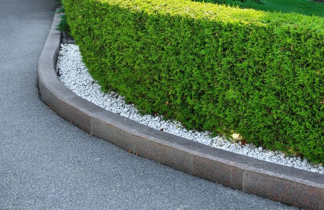 Curved concrete curb bordering a green hedge and white gravel beside a paved road
